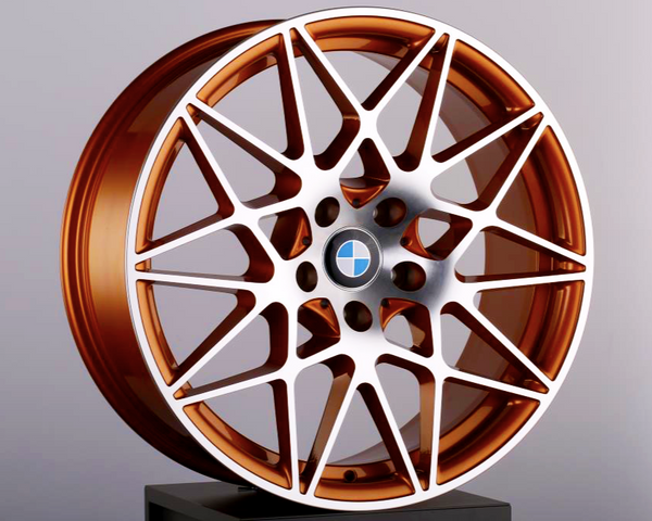 A bronze alloy wheel with intricate spoke design and a central blue and white emblem, resting on a black pedestal against a neutral gray background.