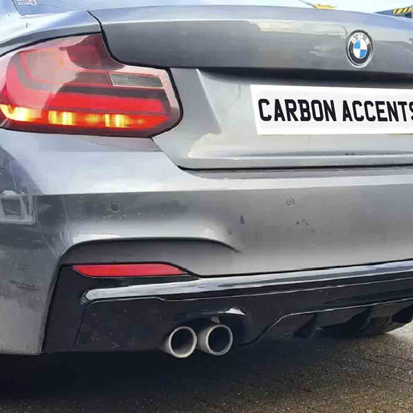 A gray BMW car is parked, displaying dual exhausts below a license plate reading "CARBON ACCENTS." It's situated on a paved surface, adjacent to a metal fence.
