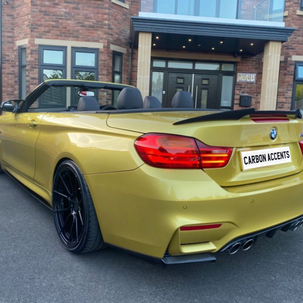 A gold convertible sports car with a visible spoiler is parked in front of a brick building. The license plate reads "CARBON ACCENTS."