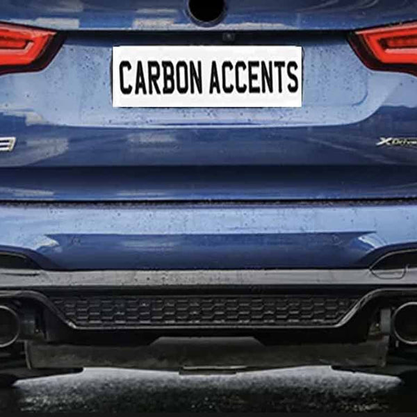 A blue car with dual exhaust pipes is parked on a street. Its license plate reads "CARBON ACCENTS." The surroundings include trees and residential buildings.
