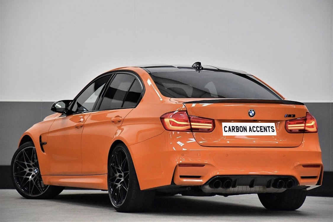Orange BMW M3, parked, features "CARBON ACCENTS" on its rear plate, displayed in a bright showroom with a grey and white wall background.