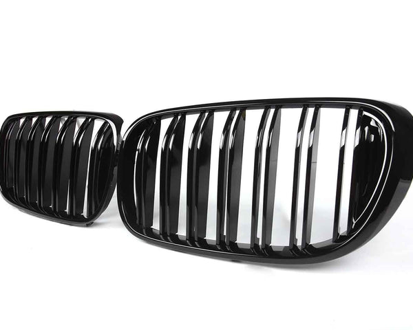 Two sleek, glossy black car grilles are positioned side by side against a plain white background, showcasing vertical slats and slightly curved oval shapes.