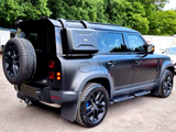 A black off-road SUV is parked on a paved area, featuring a roof rack, spare tire, and side storage box, surrounded by trees and vehicles in the background.