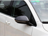 A white car features a carbon-fiber side mirror reflecting nearby trees, parked in an outdoor setting. Stickers, including a 2016 sticker, are displayed on the windshield.