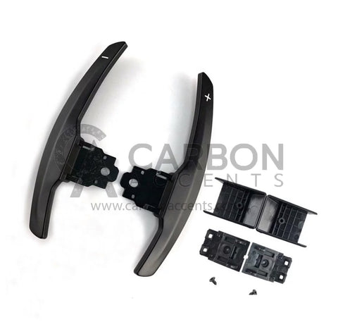 Curved paddle shifters with "+" and "-" symbols, accompanied by brackets and screws, are laid out on a white background. Text: "CARBON ACCENTS www.carbonaccents.com".