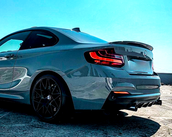 A sleek, gray sports car parked on a concrete surface, showing rear taillights and dark alloy wheels, under a clear blue sky. A warehouse is visible in the background.