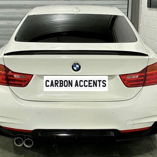 White BMW sedan with tinted windows and dual exhaust pipes is parked in a garage. The license plate reads "CARBON ACCENTS." A metallic wall and concrete floor surround the car.