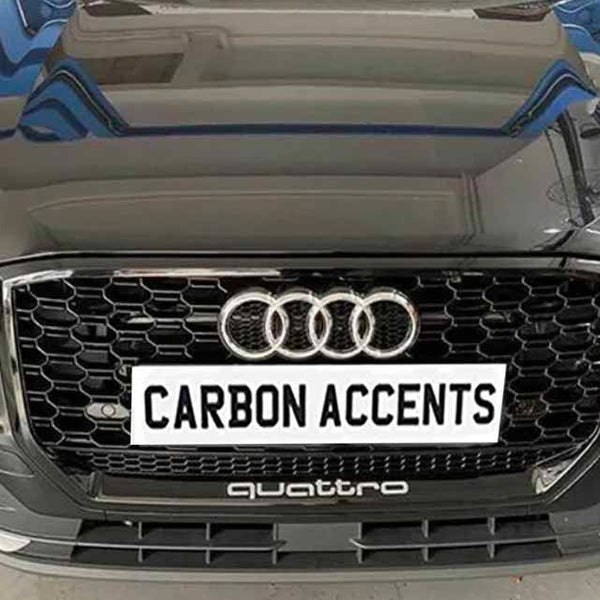 A black car's front view with a honeycomb grille and prominent logo, displaying the text "CARBON ACCENTS" on a plate, set in a garage environment with tools around.