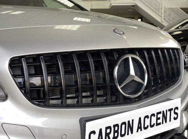 The silver car grille displays a prominent three-pointed logo, surrounded by vertical black slats in a showroom. Below, a white license plate reads "CARBON ACCENTS."