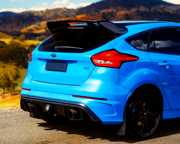 A vibrant blue sports car is parked on a roadside, featuring dual exhausts and a prominent spoiler, with scenic hills and a clear blue sky in the background.