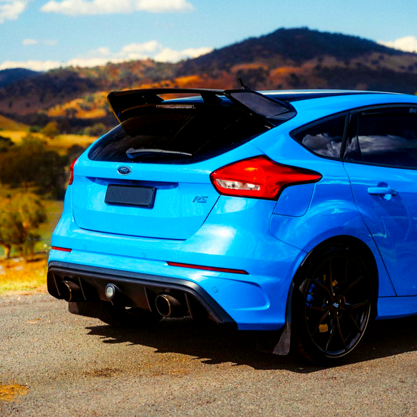 A vibrant blue sports car is parked on a roadside, featuring dual exhausts and a prominent spoiler, with scenic hills and a clear blue sky in the background.