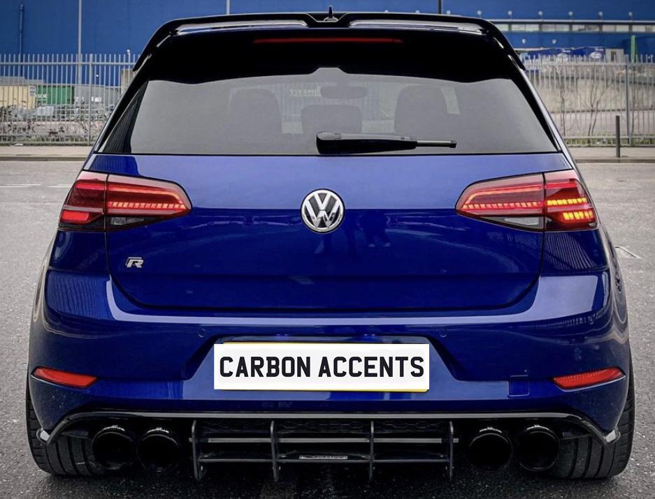 A blue Volkswagen car is parked, displaying its rear with red taillights illuminated. The license plate reads "CARBON ACCENTS," surrounded by an urban parking area with metal fencing.
