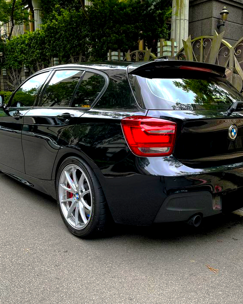 A black car is parked on a street beside ornate metal gates, with green foliage in the background. The vehicle has silver alloy wheels and a visible brand logo on the rear.