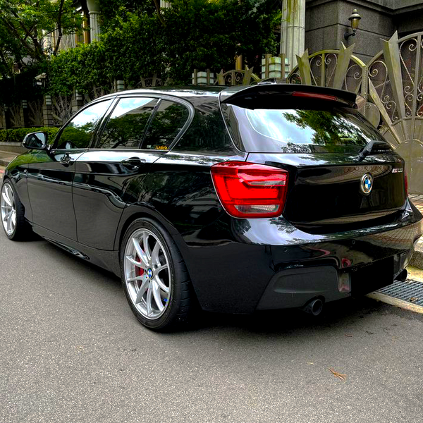 A black car is parked on a street beside ornate metal gates, with green foliage in the background. The vehicle has silver alloy wheels and a visible brand logo on the rear.