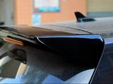 A black car roof spoiler with a reflective finish is mounted on a vehicle, parked in an urban setting. A building's blurred exterior is visible in the background.