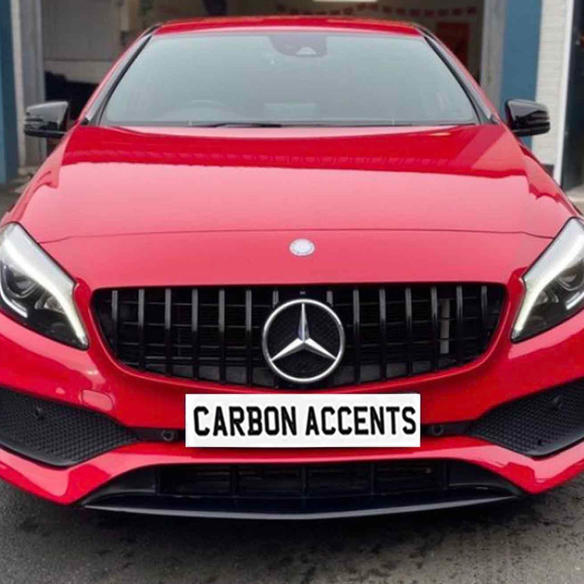 A red car featuring a prominent front grille and sleek headlights is parked outside a garage. The license plate reads "CARBON ACCENTS."