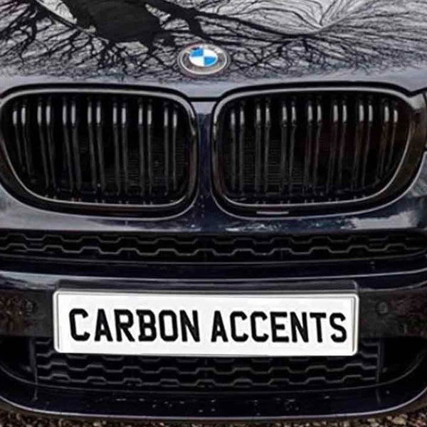 A black car displays "CARBON ACCENTS" on its front license plate. The headlights are on, and tree branches are reflected on the hood, parked on a gravel surface.