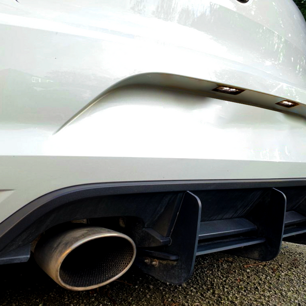A white car's rear with dual exhaust pipes and a black diffuser sits on a gravel driveway, bordered by greenery, under a clear sky.