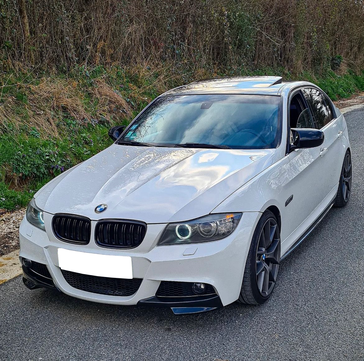 A white car with a sleek design is parked on a road, surrounded by grass and bushes. The car features distinctive headlights and a sunroof.