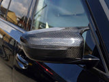 Carbon fiber car side mirror reflecting the sunlight, attached to a sleek black vehicle. The background shows blurred buildings and a clear sky.