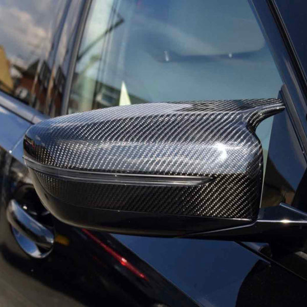 Carbon fiber car side mirror reflecting the sunlight, attached to a sleek black vehicle. The background shows blurred buildings and a clear sky.