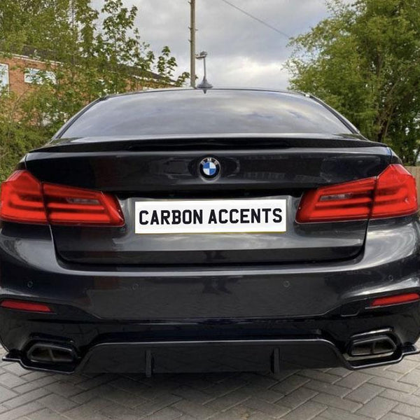 A black BMW car is parked with its back facing the viewer, displaying a license plate reading "CARBON ACCENTS," in a driveway with trees and a brick building nearby.