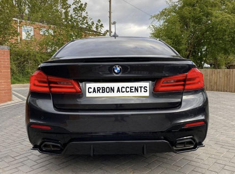 A black BMW car is parked with its back facing the viewer, displaying a license plate reading "CARBON ACCENTS," in a driveway with trees and a brick building nearby.