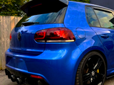 A blue car with tinted taillights and a prominent spoiler is parked on a gravel surface, next to a wooden fence and greenery, under a partly cloudy sky.