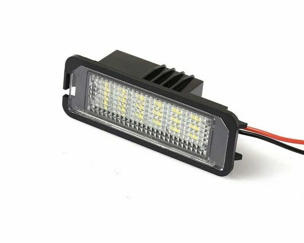 A rectangular LED light is resting on a white surface, featuring multiple small lights behind a textured lens, with red and black wires extending from its back.