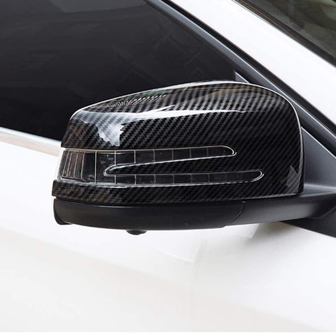A car's side mirror with a carbon fiber design cover, featuring integrated indicator lights, mounted on a white vehicle in an outdoor setting.