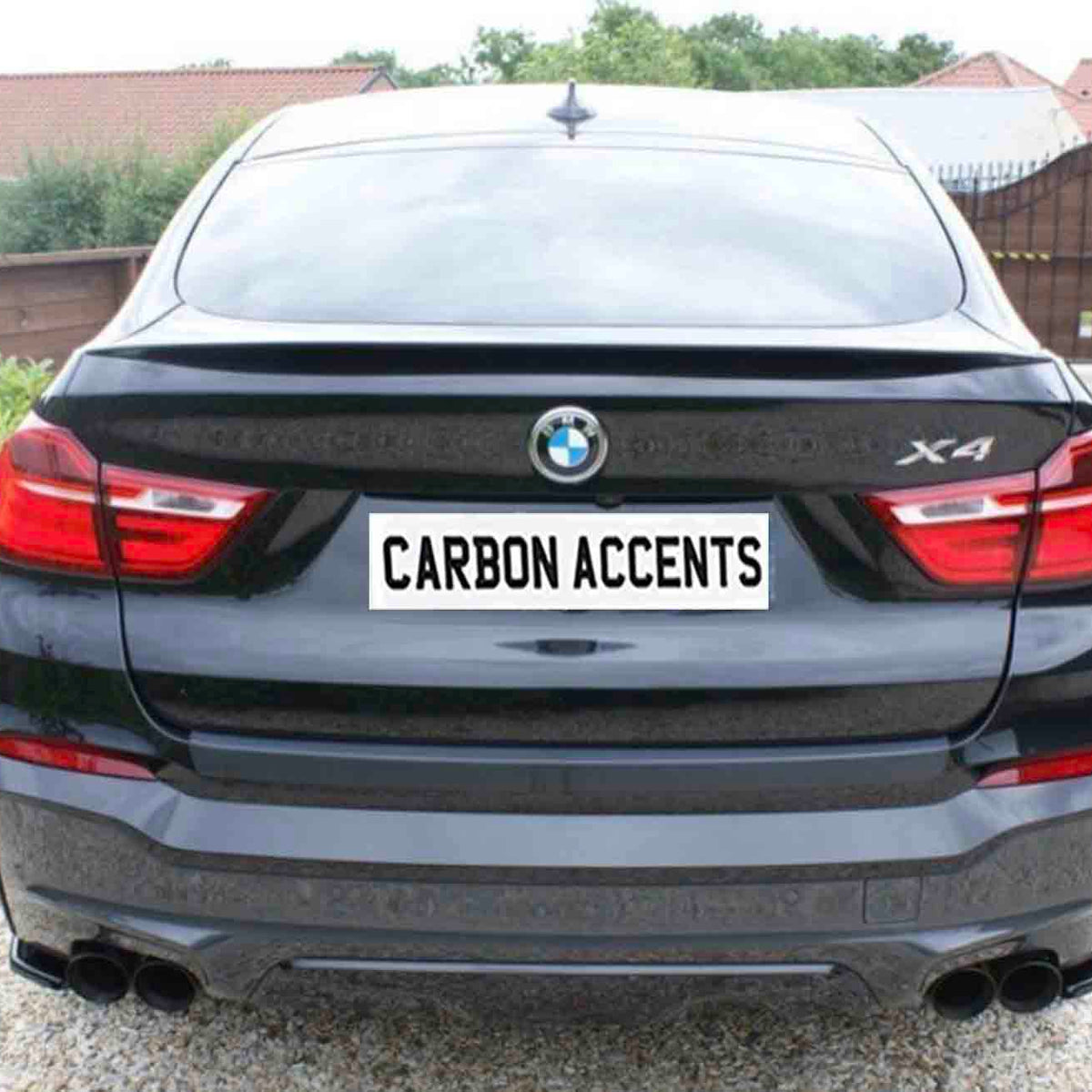 A black BMW X4 with visible taillights is parked on gravel. The license plate reads "CARBON ACCENTS." Trees and a fence form the background environment.
