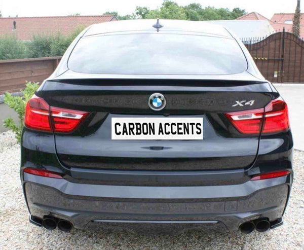 A black BMW X4 with visible taillights is parked on gravel. The license plate reads "CARBON ACCENTS." Trees and a fence form the background environment.