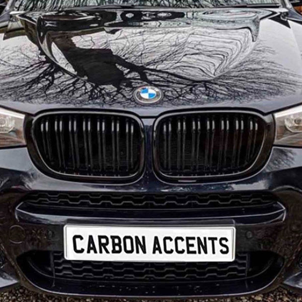 A black car displays headlights in a driveway, with a front plate reading "CARBON ACCENTS." Trees are reflected on the hood, and a wooden fence lines the background.
