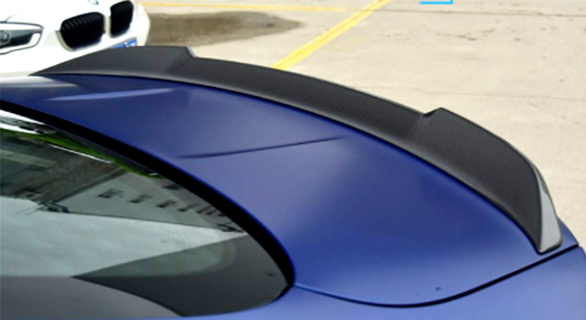 A blue car features a black spoiler, parked on a concrete surface. Another white car is partially visible in the background, highlighting a parking lot setting.