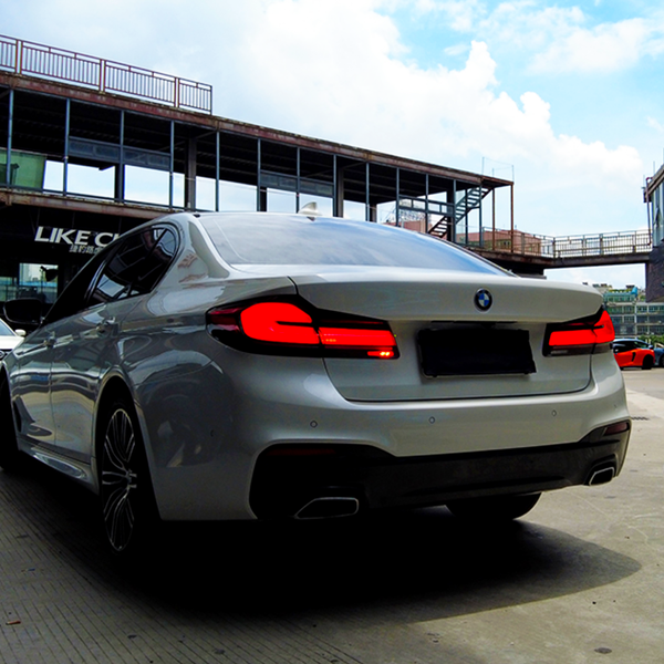 A white BMW car is parked on a concrete surface with its taillights on, surrounded by a building complex. Another car and signs, including "LIKE C," are visible nearby.