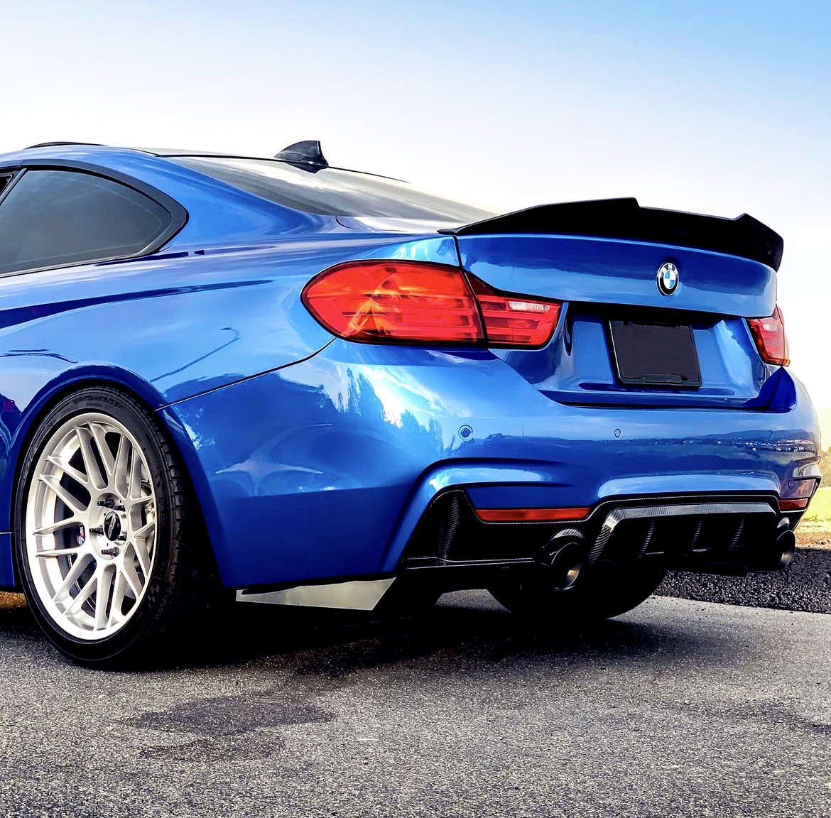 A shiny blue sports car with a spoiler is parked on a paved surface. Its red taillights are illuminated, and the background shows a clear blue sky.