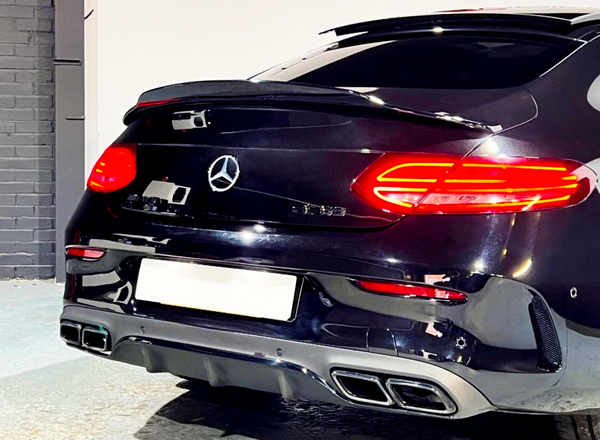 A black car parked indoors, displaying illuminated taillights. The Mercedes-Benz emblem is centered on the trunk. The garage background features grey and white walls.
