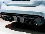 A car's rear features four exhaust pipes integrated into a carbon-fiber diffuser, emitting a sleek, high-performance appearance. The setting is well-lit, highlighting the vehicle's smooth white exterior.