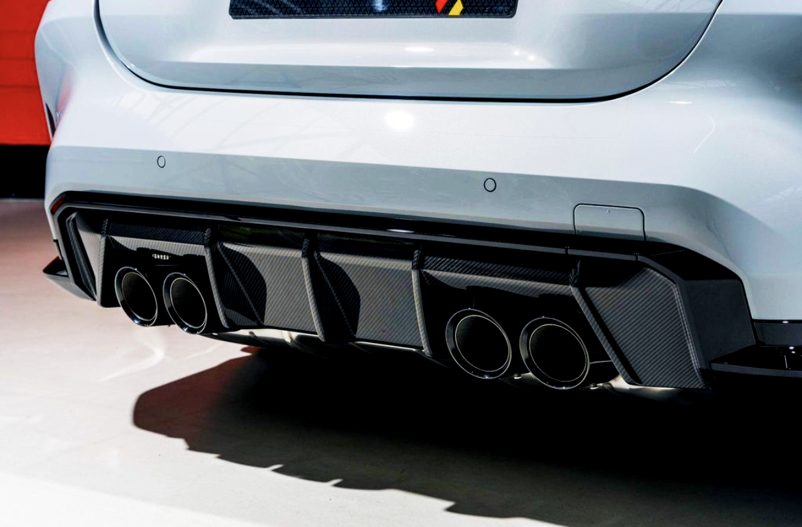 A car's rear features four exhaust pipes integrated into a carbon-fiber diffuser, emitting a sleek, high-performance appearance. The setting is well-lit, highlighting the vehicle's smooth white exterior.