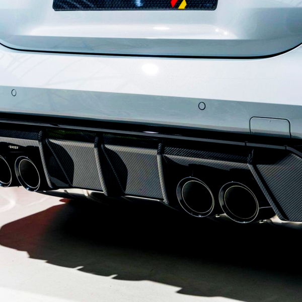A car's rear features four exhaust pipes integrated into a carbon-fiber diffuser, emitting a sleek, high-performance appearance. The setting is well-lit, highlighting the vehicle's smooth white exterior.