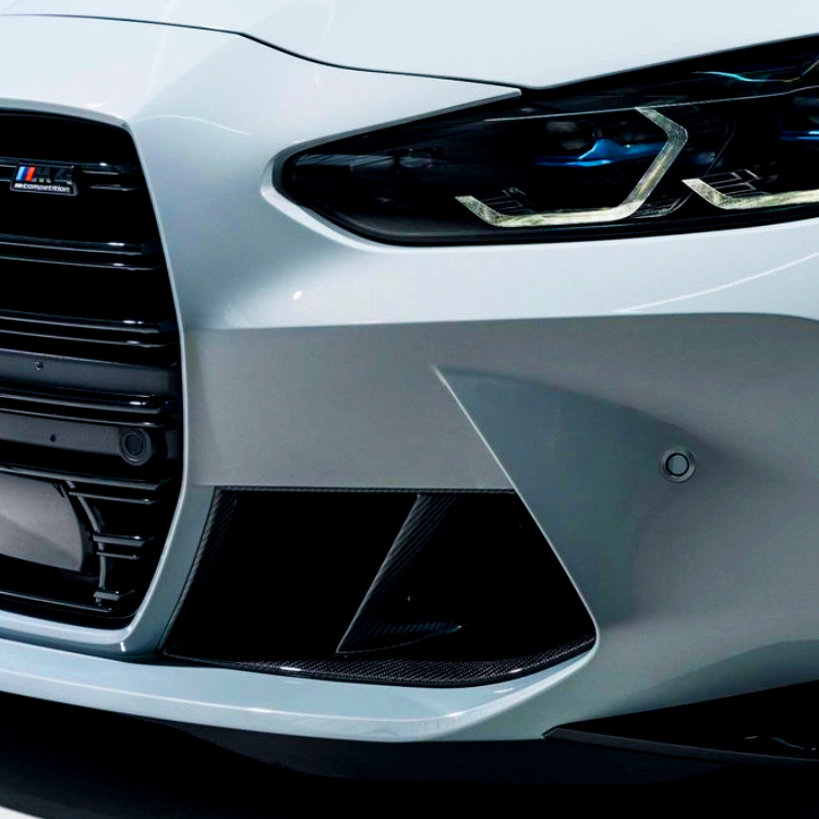 The image shows the front end of a white car with sleek, angular headlights and a prominent grille. "M Competition" logo is visible, indicating it's a BMW model.
