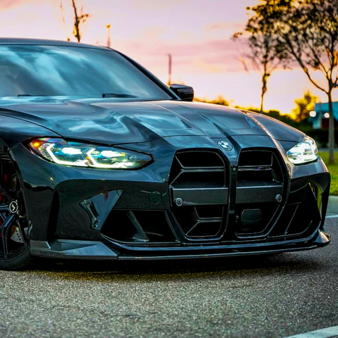 A sleek, glossy black sports car is parked on a paved road at sunset. Its prominent grille and illuminated headlights are emphasized against a backdrop of trees and a colorful sky.