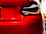 A red car's rear end, featuring illuminated taillights with an intricate design, is parked inside a garage. Reflections of lights appear on the glossy surface of the vehicle.