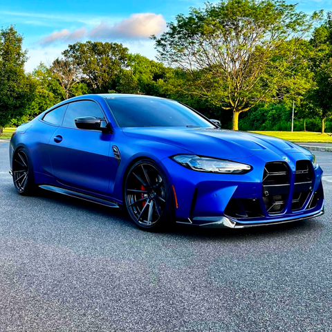 A blue sports car is parked on a paved road, surrounded by lush green trees and a clear blue sky in the background.