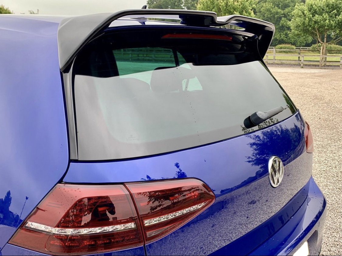 A blue Volkswagen hatchback parked in a gravel area features a rear wiper and tinted windows. Nearby, lush green trees and a wooden fence complete the serene setting.