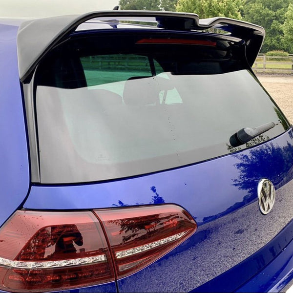 A blue Volkswagen hatchback parked in a gravel area features a rear wiper and tinted windows. Nearby, lush green trees and a wooden fence complete the serene setting.