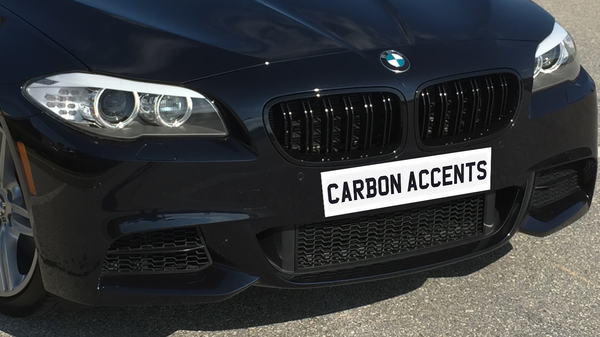 A close-up of a black BMW car's front grille and headlights, parked on a sunlit road, displays the text "CARBON ACCENTS" on the license plate area.