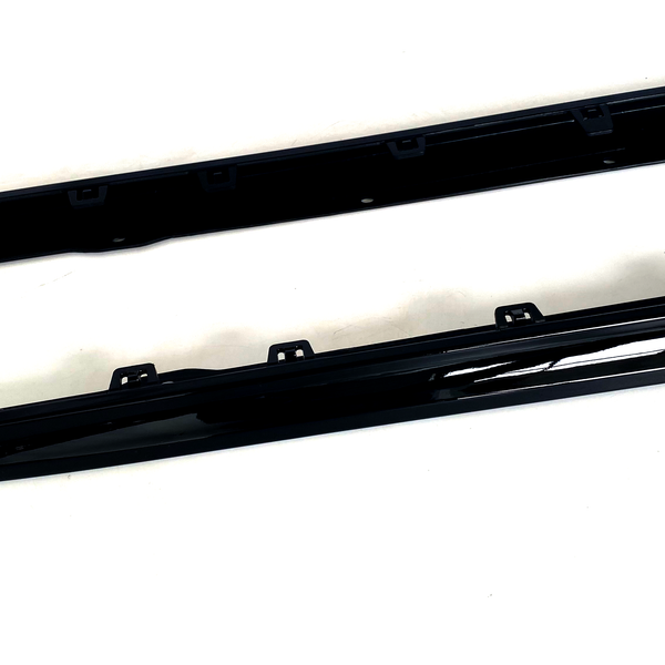 Two black car side skirts are resting on a white surface, featuring several mounting brackets along their length for attachment.