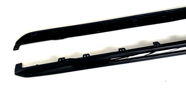 Two black car side skirts are resting on a white surface, featuring several mounting brackets along their length for attachment.