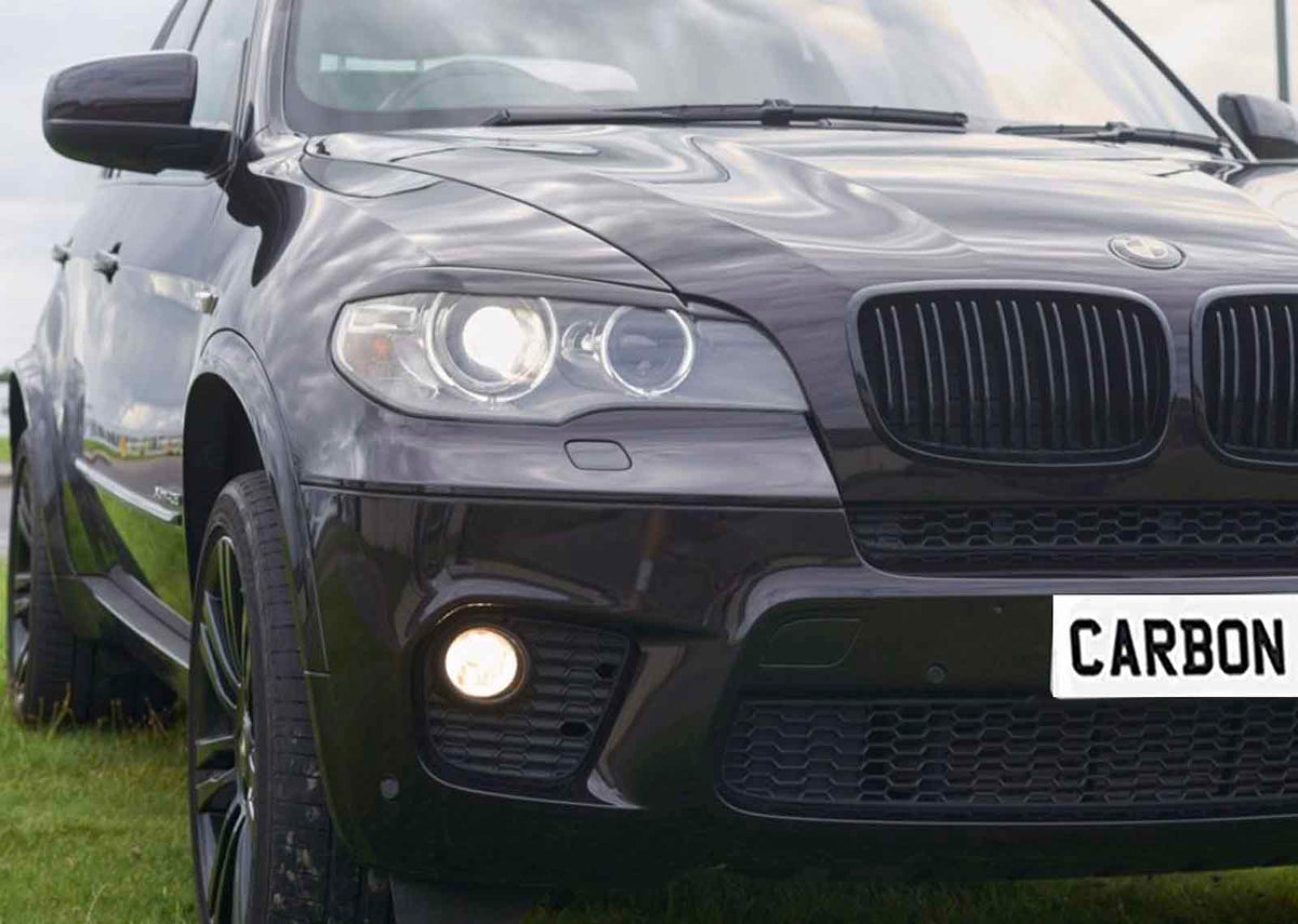 A dark SUV with glowing headlights is parked on grass, featuring reflective paint and a front license plate reading "CARBON." The sky is cloudy.